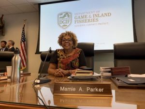 Dr. Mamie Parker sits at a large table with a nameplate. She is seated in an executive chair in front of a screen that says Dept of Game & Inland Fisheries